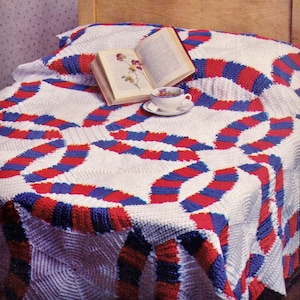 Könnte beinhalten: Eine weiße Häkeldecke mit einem rot-weiß-blauen Muster. Die Decke ist gefaltet und über ein Bett gelegt, auf dem ein Buch und eine Tasse Tee liegen.