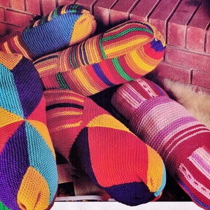 May include: A set of three colourful knitted cushions with geometric patterns in shades of red, yellow, blue, green, and purple. The cushions are arranged in a pile, with the largest cushion on the bottom and the smallest cushion on top.