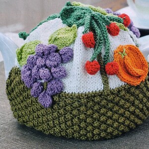 Può includere: Una coperta per teiera lavorata a maglia con sfondo bianco, ornata da foglie verdi, uva viola, ciliegie rosse e un frutto arancione. Il bordo inferiore è verde oliva strutturato. Per una teiera bianca.