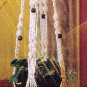 May include: White macrame plant hanger with brown wooden beads, holding a black pot with green plants.