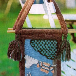 May include: A brown knitted bag with a blue and green knitted design. The bag has two brown tassels and a wooden handle. The bag is hanging on a white chair.