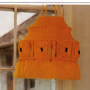 May include: A handmade, orange macrame lampshade with a tiered design. The lampshade features a series of geometric patterns and is adorned with small, black beads. It hangs from a window, showcasing its unique, handcrafted aesthetic.