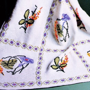 May include: White crocheted blanket with a repeating pattern of a green and purple bird, a dragonfly with orange and yellow wings, and a green and orange flower. The blanket has a border of purple and green flowers.
