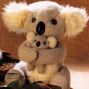 Puede incluir: Un koala de crochet gris que sostiene un koala de crochet gris más pequeño. Ambos koalas tienen pelo blanco en las orejas y están sentados sobre una superficie marrón.