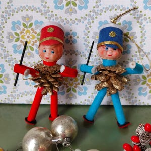 May include: Two vintage Christmas ornaments shaped like people wearing red and blue felt uniforms with pine cone accents. The ornaments have black felt sticks and gold accents.