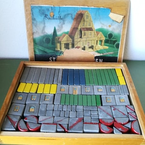 Può includere: Una scatola di legno piena di blocchi di legno colorati. La scatola ha un coperchio con un'immagine di una casa e il testo "Bauh". I blocchi sono di varie forme e dimensioni, tra cui quadrati, rettangoli e triangoli. I blocchi sono grigi, verdi, blu, gialli e rossi.