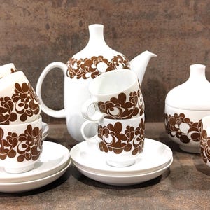 Op de afbeelding: Een wit keramisch theeservies met een bruin bloemmotief. De set bevat een theepot, suikerpot, melkkan, kopjes en schotels. De kopjes zijn opgestapeld en de set is geplaatst op een bruin oppervlak.