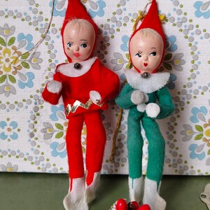 Könnte beinhalten: Zwei vintage Weihnachts-Elfen-Ornamente aus Filz. Ein Elf trägt ein rotes Kostüm mit goldenen Verzierungen und der andere ein grünes Kostüm. Beide Elfen haben weiße Filzstiefel und weiße Filzkragen.