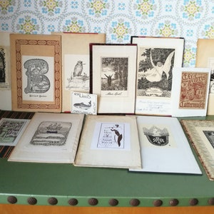 Könnte beinhalten: Eine Sammlung von 15 gerahmten Schwarzweißdrucken, einige mit Text, auf einem grünen Tisch mit braunem Holzsockel ausgestellt. Die Drucke sind alle unterschiedlich groß und zeigen verschiedene Bilder, darunter einen Baum, ein Gebäude, ein Schiff, einen Vogel und eine Person.