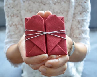 Puede incluir: Una caja de regalo rectangular envuelta en tela de color rojo intenso con un diseño de corazón de origami plegado en la parte frontal. El paquete está asegurado con un cordel metálico plateado fino envuelto horizontalmente alrededor de la tela.