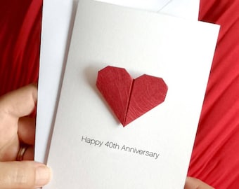 May include: A white greeting card with a red origami heart and a matching white envelope. The card reads "Happy 40th Anniversary" in a simple, elegant font. The background is a solid red fabric.