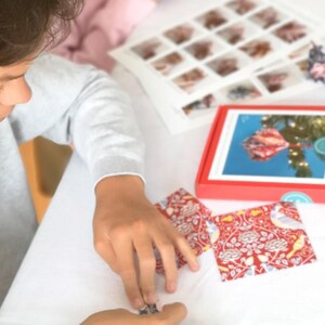May include: A person is working on a craft project. The image shows a red box with a picture of a Christmas ornament, and two red patterned cards with birds and flowers. The cards are on a white surface.