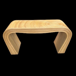 May include: A light tan console table constructed from stacked layers of wood, featuring a smooth, continuous curved design that forms the legs and top. The surface displays a prominent circular grain pattern on the right side of the tabletop.
