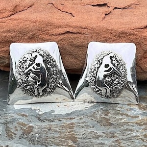 May include: A pair of silver cufflinks with a rectangular shape and a textured, circular design in the center. The design features a silhouetted figure. The cufflinks are displayed on a gray and brown surface.
