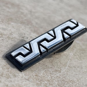 Könnte beinhalten: Ein silberner Krawattenclip mit geometrischem Design. Das Design besteht aus einer Reihe ineinandergreifender Dreiecke, die ein sich wiederholendes Muster bilden.