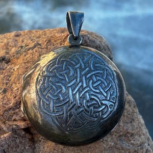 Puede incluir: Un colgante de medallón redondo de metal plateado con un intrincado diseño de nudos celtas en relieve en la parte frontal. El colgante incluye una resistente anilla de metal pulido en la parte superior para sujetarlo a una cadena o collar.