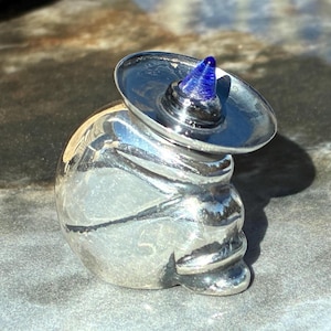 May include: A silver-colored skull-shaped bottle stopper with a small sombrero. The sombrero has a blue glass cone-shaped top. The stopper is made of a shiny, reflective metal.
