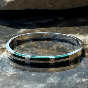 May include: A silver-toned hinged bangle bracelet featuring inlaid rectangular segments of green malachite stone. The interior of the metal band is engraved with the text "MEXICO" and "TM-228". The bracelet has a smooth, polished finish.