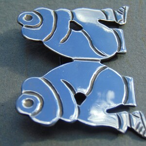 May include: A silver brooch with a stylized design of two figures, possibly fish, facing each other. The figures are detailed with lines and curves, and the brooch has a polished finish.