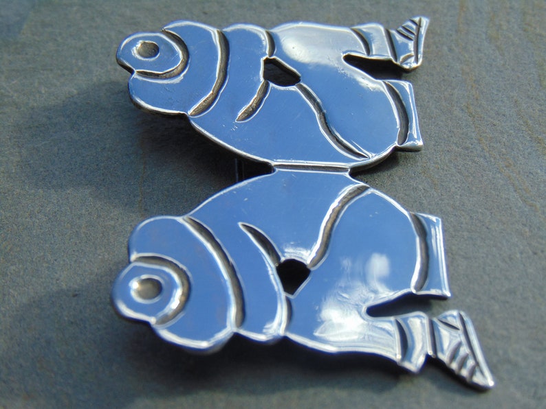 May include: A silver brooch with a stylized design of two figures, possibly fish, facing each other. The figures are detailed with lines and curves, and the brooch has a polished finish.