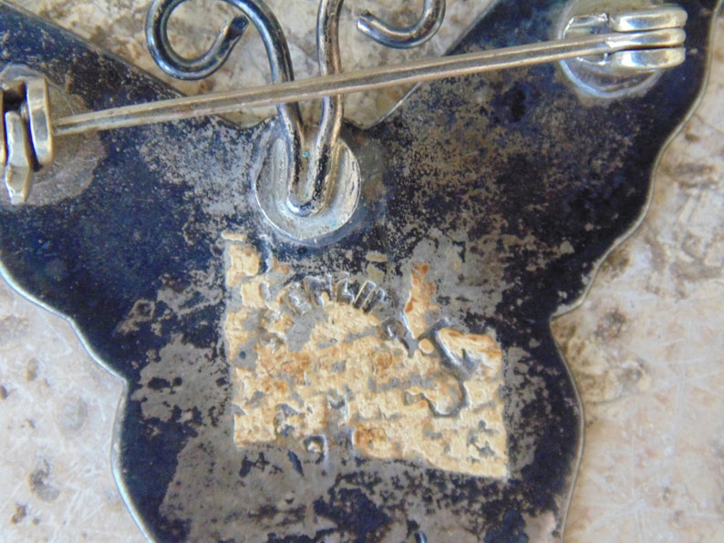 May include: The back of a silver butterfly brooch with a pin and a maker's mark. The mark is a stylized letter 'S' with a swirl design.