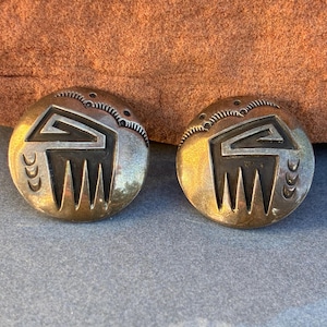 以下が含まれることがあります： 南西部のデザインが施された、丸い銀色のイヤリングのペア。各イヤリングは、一連の垂直線の上に中央の幾何学的な形状があり、端に装飾的な要素があります。イヤリングは灰色の表面に表示されています。