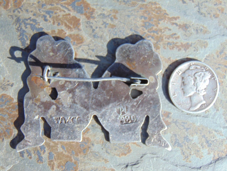 May include: Silver tone pin with two figures holding hands. The back of the pin has the markings "TAVCC" and "920".
