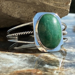 May include: A silver cuff bracelet featuring a large, oval, green gemstone. The gemstone is set in a rectangular silver frame. The bracelet has a twisted rope design on the sides and a simple, curved band.
