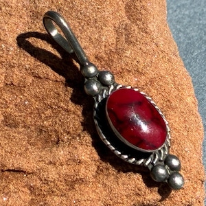 May include: A silver pendant with an oval, deep red stone. The stone is set in a decorative silver frame with a twisted rope design. The pendant has a loop at the top for a chain.