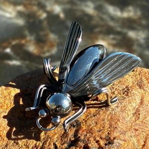 Puede incluir: Un broche con forma de insecto que presenta un cuerpo ovalado oscuro, liso y pulido, una cabeza redonda y alas alargadas y estriadas. La pieza está construida con alambre de metal plateado brillante y componentes de piedra oscura reflectante.
