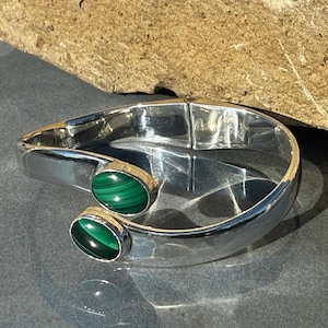 May include: A polished silver hinged cuff bracelet featuring two oval-shaped malachite gemstones. The bracelet has a smooth, reflective metallic finish and an open-ended design where the two green, banded malachite stones face each other.