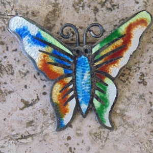 May include: A colorful butterfly brooch with enamel detailing in shades of blue, green, orange, and white. The butterfly has black lines on its wings and a silver metal body.