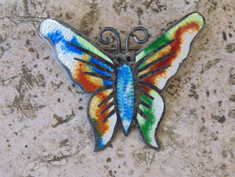 May include: A colorful butterfly brooch with enamel detailing in shades of blue, green, orange, and white. The butterfly has black lines on its wings and a silver metal body.