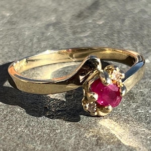 May include: A gold ring with a red ruby gemstone surrounded by smaller diamonds. The ring has a simple band and a delicate design.