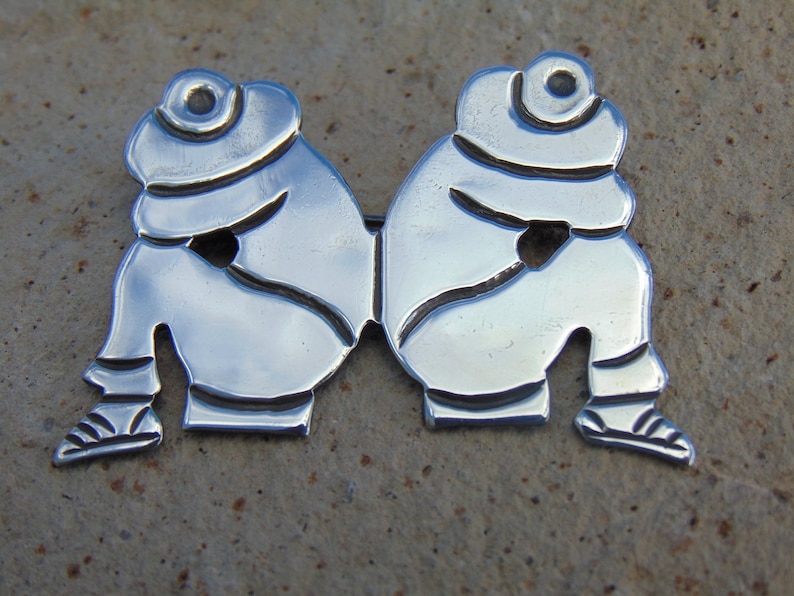 May include: A silver brooch depicting two figures sitting back to back, each wearing a hat. The figures are stylized and have a simple, minimalist design.