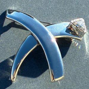 Op de afbeelding: Zilverkleurige broche met een gepolijste afwerking, bestaande uit twee elkaar kruisende gebogen staven en een facetgeslepen, diamantvormig accent. De broche heeft een reflecterend oppervlak en een goudkleurige rand.