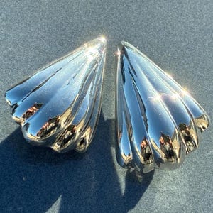 May include: A pair of shiny silver-colored earrings. Each earring has a fan-like shape with multiple curved ridges, reflecting light. The earrings are displayed on a dark gray surface, with sunlight creating bright highlights.
