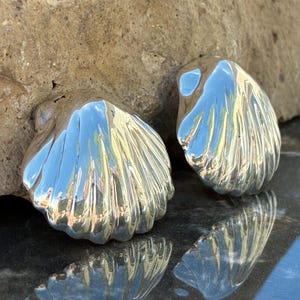 May include: A pair of polished silver-toned earrings shaped like fan-style seashells. Each earring features a smooth, reflective surface with deep, vertical ridges radiating from the base to the scalloped edge, creating a high-shine metallic finish.