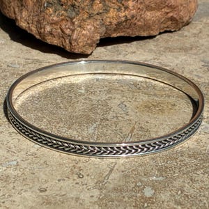 Op de afbeelding: Een zilverkleurige armband met een gevlochten ontwerp rond de rand. De armband is cirkelvormig en lijkt van metaal te zijn gemaakt. De achtergrond bevat een rots en een getextureerd oppervlak.