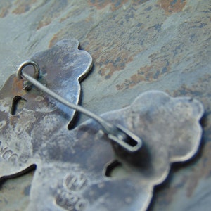 May include: A silver-toned pin with two stylized figures. The back of the pin has the markings "T.M.C.C." and "M.J.S.C."