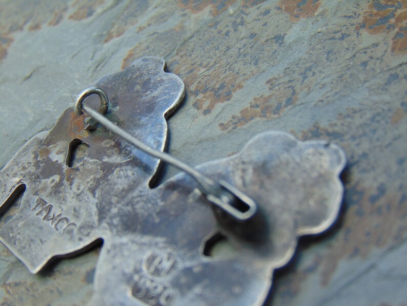 May include: A silver-toned pin with two stylized figures. The back of the pin has the markings "T.M.C.C." and "M.J.S.C."