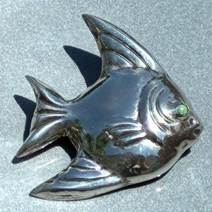 Puede incluir: Broche plateado con forma de pez brillante. El broche presenta detalles finos, incluyendo aletas y cola. Un pequeño ojo verde redondo está incrustado. La superficie metálica refleja la luz, sugiriendo un pulido.