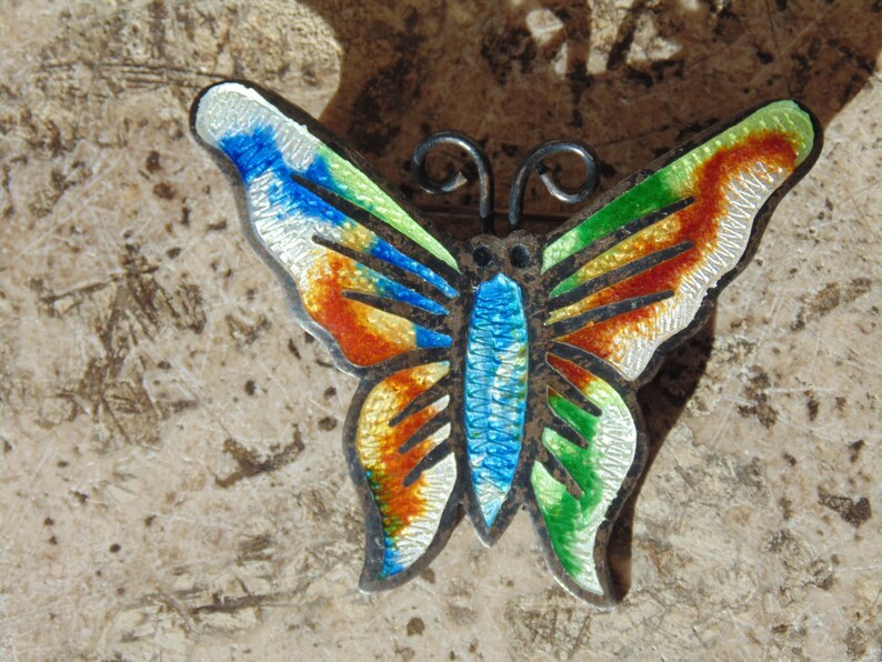 May include: A silver butterfly brooch with colorful enamel detailing. The wings are decorated with green, blue, red, and white enamel. The butterfly is a popular motif in jewelry.