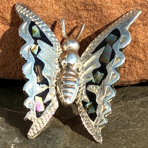 May include: A silver-toned butterfly brooch with intricate detailing. The wings feature black inlays with iridescent accents, and the body has a ribbed texture. The brooch is designed with delicate antennae and wing edges.