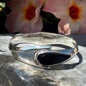 May include: A polished silver-tone hinged bangle bracelet featuring a smooth, teardrop-shaped black inlay at the center. The metal surface is highly reflective, showcasing a sleek, curved design that tapers toward the focal point of the black stone.