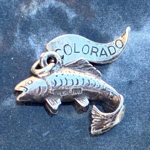 Puede incluir: Un colgante plateado con un diseño detallado de pez y la palabra "COLORADO" encima. El colgante probablemente está hecho de plata de ley y es un amuleto o una pequeña joya.