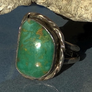 May include: A silver ring with a large, oval turquoise stone. The stone is a vibrant green with brown and gold inclusions. The ring has a twisted rope design around the stone and a split shank.