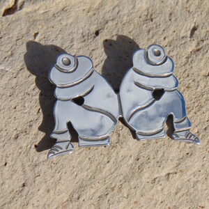 May include: Silver brooch depicting two figures wearing hats and sitting side-by-side.