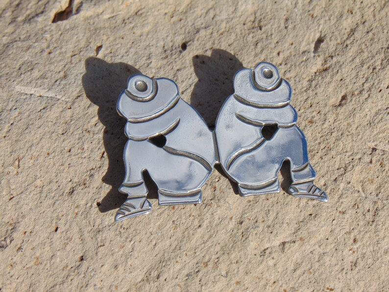 May include: Silver brooch depicting two figures wearing hats and sitting side-by-side.