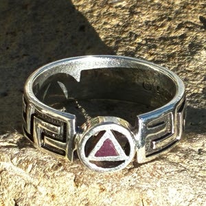 Puede incluir: Un anillo de plata con un emblema circular central que contiene un triángulo granate. La banda está decorada con un patrón geométrico griego repetitivo a ambos lados del círculo. El metal tiene un acabado pulido con zonas oscuras.